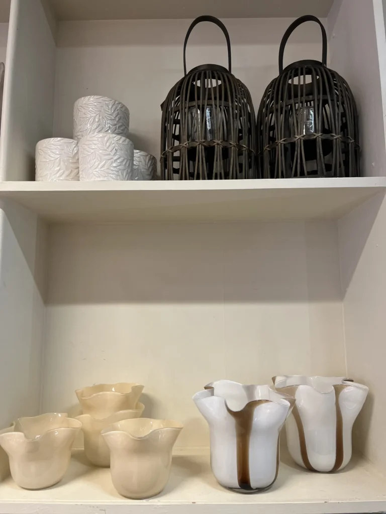 Keramiska ljusstakar, dekorativa korglyktor i brunt och vita koppar med textur är placerade på två vita hyllor.