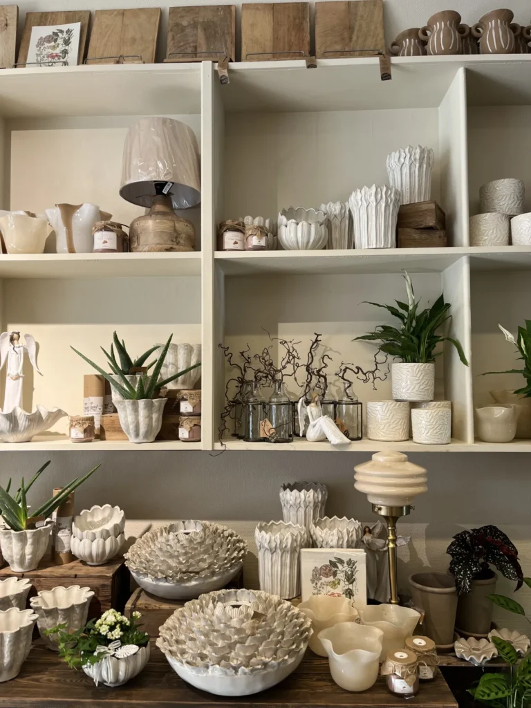 På hyllor och ett bord står olika vita keramikkrukor, skålar och vaser, vissa med gröna växter i, i en prydlig miljö med dekorativa föremål.
