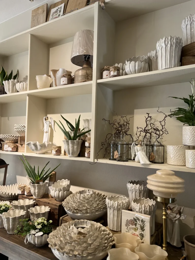 Vita keramikvaser, skålar och krukor med växter och prydnadsföremål är placerade på hyllor och ett träbord mot en beige vägg.