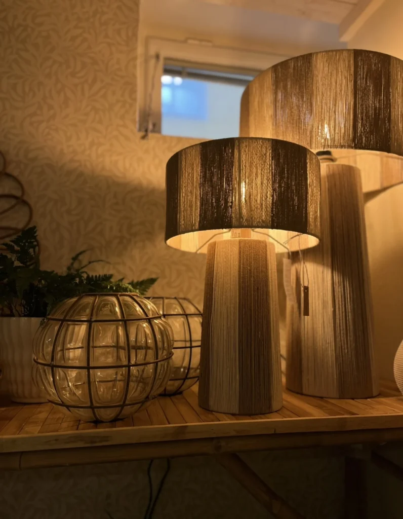 Två beige bordslampor med cylindriska fötter och texturerade skärmar står på en träyta nära ett dekorativt föremål i burglas och en krukväxt, med ett fönster i bakgrunden.