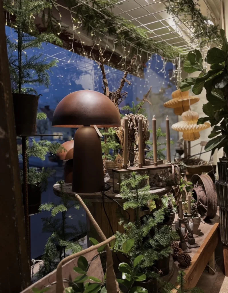Inomhusutställning med brun svampformad lampa, olika krukväxter, ljusslingor och papperslyktor, mot ett fönster som visar en mörk himmel utanför.