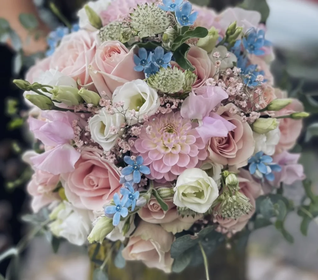 En bukett med pastellfärgade blommor, inklusive ljusrosa rosor, vita lisianthus, blå förgätmigej och grönska, arrangerade i en tät, rundad form.