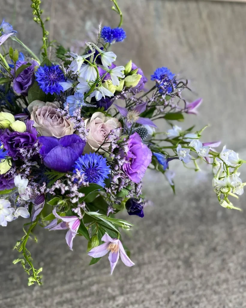 En bukett lila, blå och vita blommor med grönt bladverk mot en grå betongbakgrund.