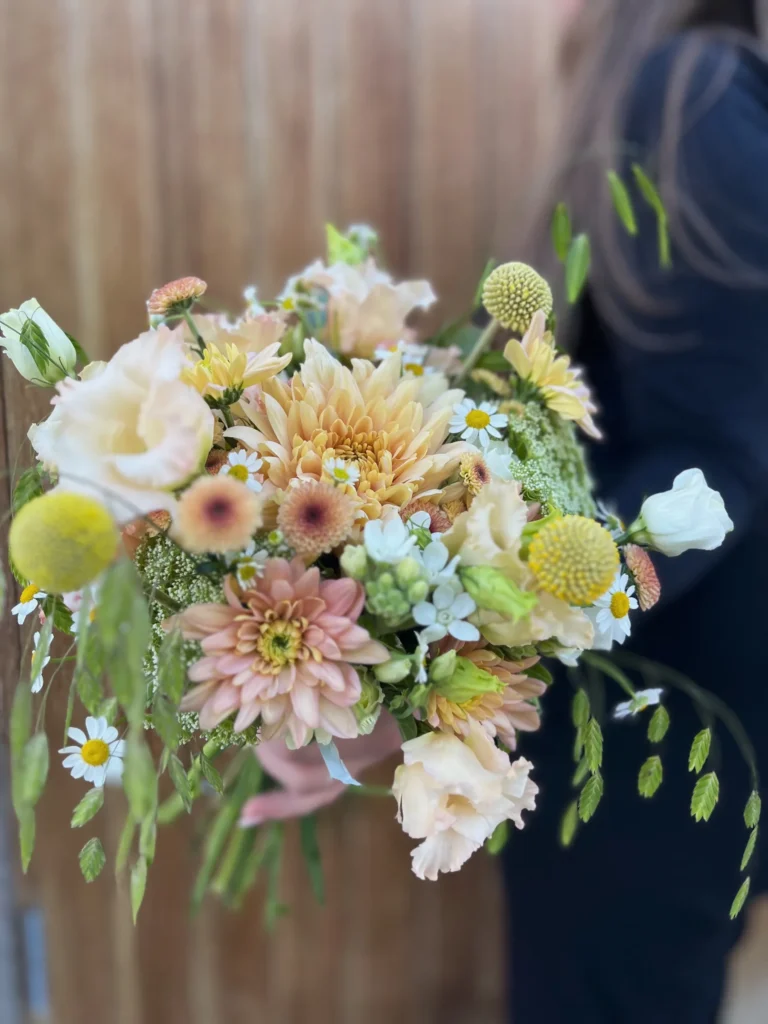 En mörkklädd person håller i en blandad blombukett med gula, persikofärgade och vita blommor och gröna blad mot en suddig brun bakgrund.