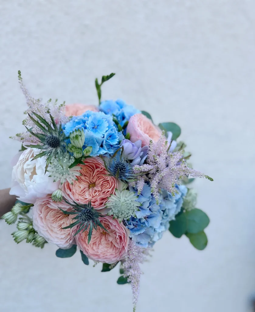 En bukett med persikofärgade rosor, blå hortensior, vita ranunkler och diverse grönska mot en enfärgad, ljus bakgrund.