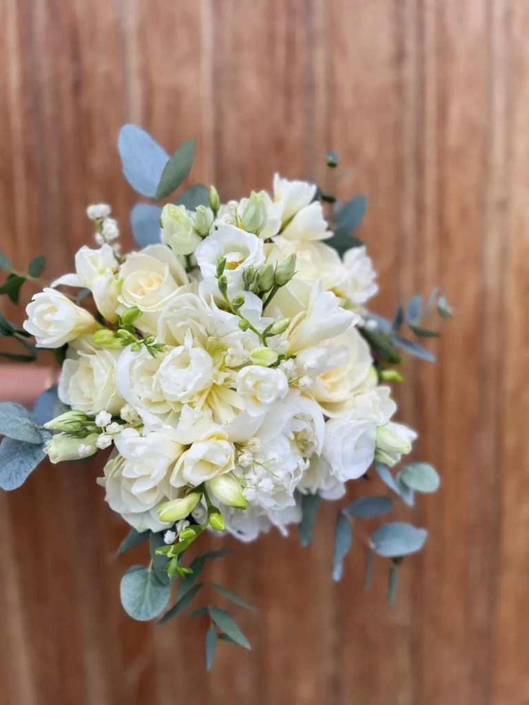 En bukett vita och gräddfärgade rosor med gröna eukalyptusblad mot en träbakgrund.