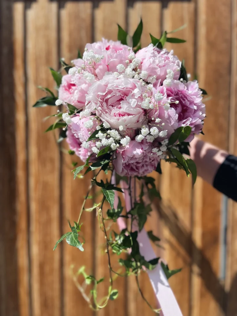 En hand håller en bukett med rosa pioner, babys breath och grönska, med efterföljande vinrankor och ett rosa band, framför en träbakgrund.