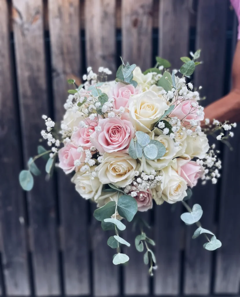 En bukett av vita och ljusrosa rosor med babys breath och eukalyptusblad, framför ett trästaket.