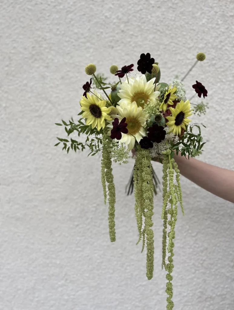En hand håller en bukett med solrosor, mörkt rödbruna blommor, vita blommor, gröna blad och gröna amaranthus mot en enfärgad, ljus vägg.