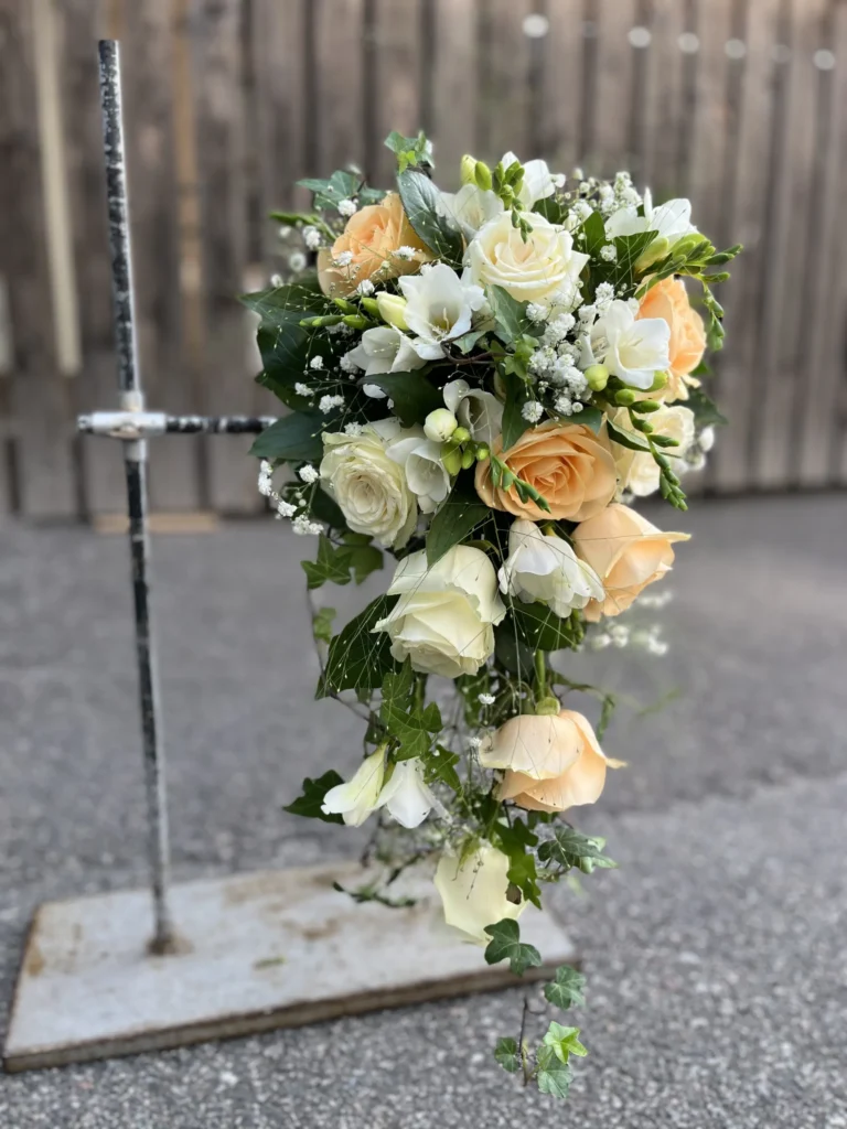 En kaskadbukett av vita och persikofärgade rosor med grönska och babys breath, placerad på ett metallstativ utomhus mot ett trästaket.