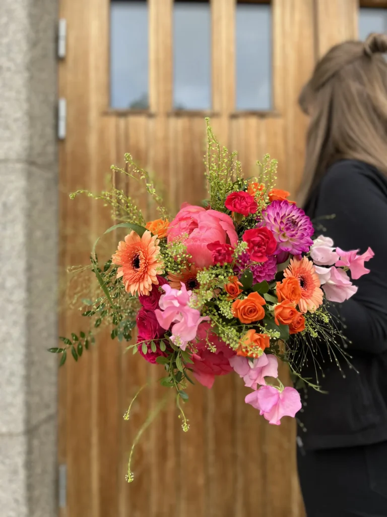 Person som håller i en livlig bukett med blandade blommor, bland annat rosa pioner, orange gerbera och rosor, framför en trädörr.