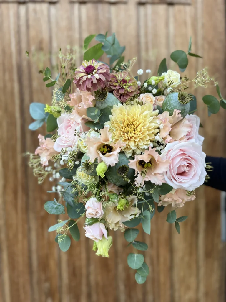 En hand håller en bukett pastellfärgade blommor, inklusive rosor, dahlior, lisianthus och diverse grönska, mot en träbakgrund.