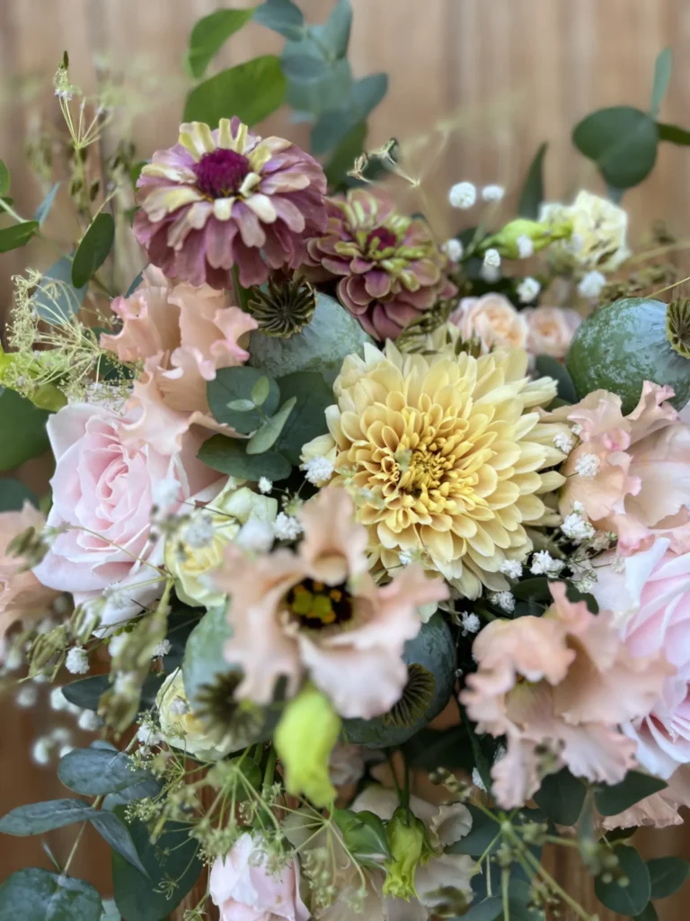 En bukett med ljusgula, ljusrosa och lila blommor med gröna blad och knoppar i ett naturligt, löst arrangemang.