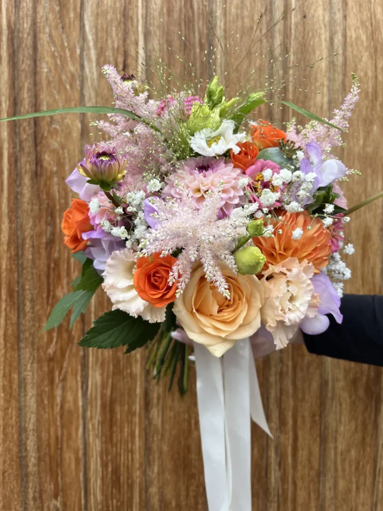 En hand håller en bukett med blandade blommor, inklusive orangefärgade rosor, persikofärgade nejlikor och lavendelblommor, knutna med ett vitt band, framför en träbakgrund.
