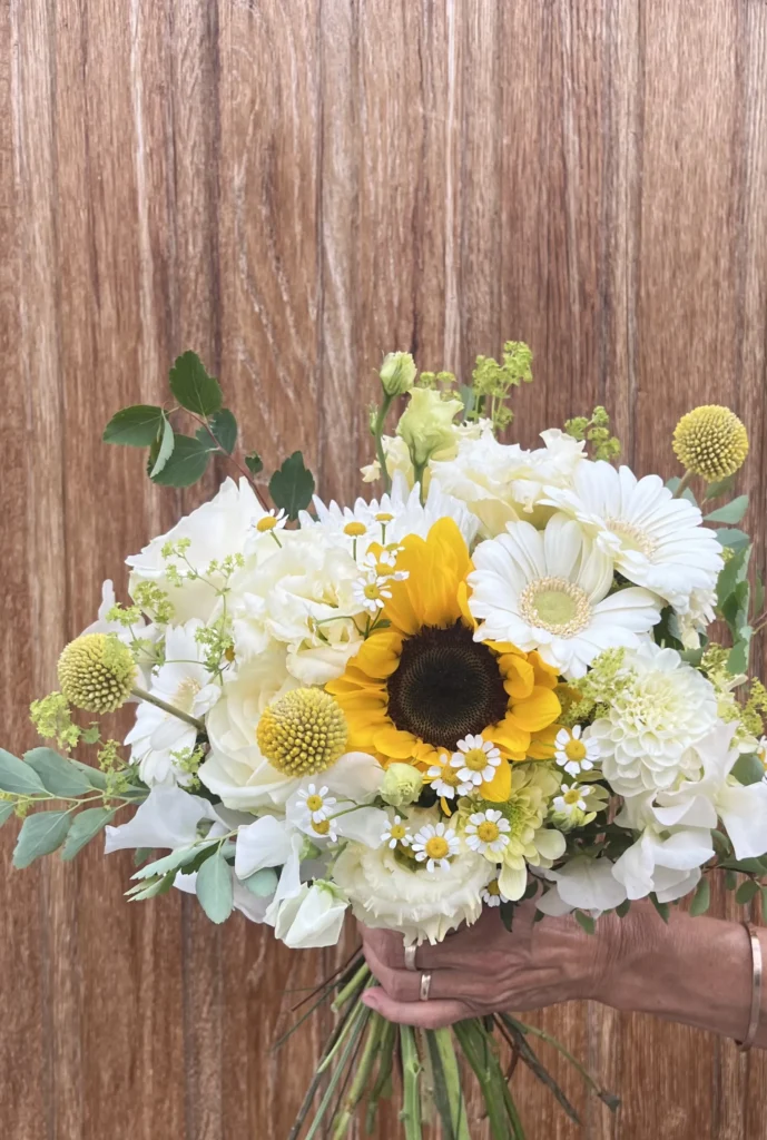 En hand håller i en blombukett med solrosor, vita rosor, prästkragar och grönt, mot en bakgrund av trä.