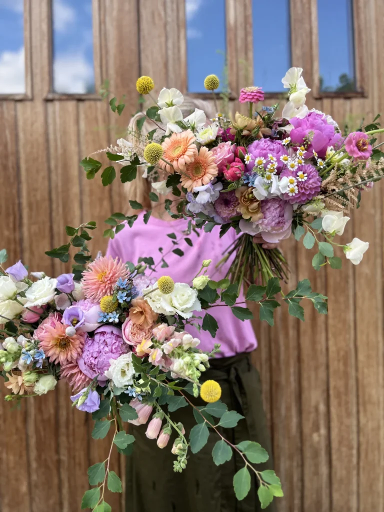 En person i rosa skjorta håller två stora, färgglada blomsterarrangemang framför en trädörr.
