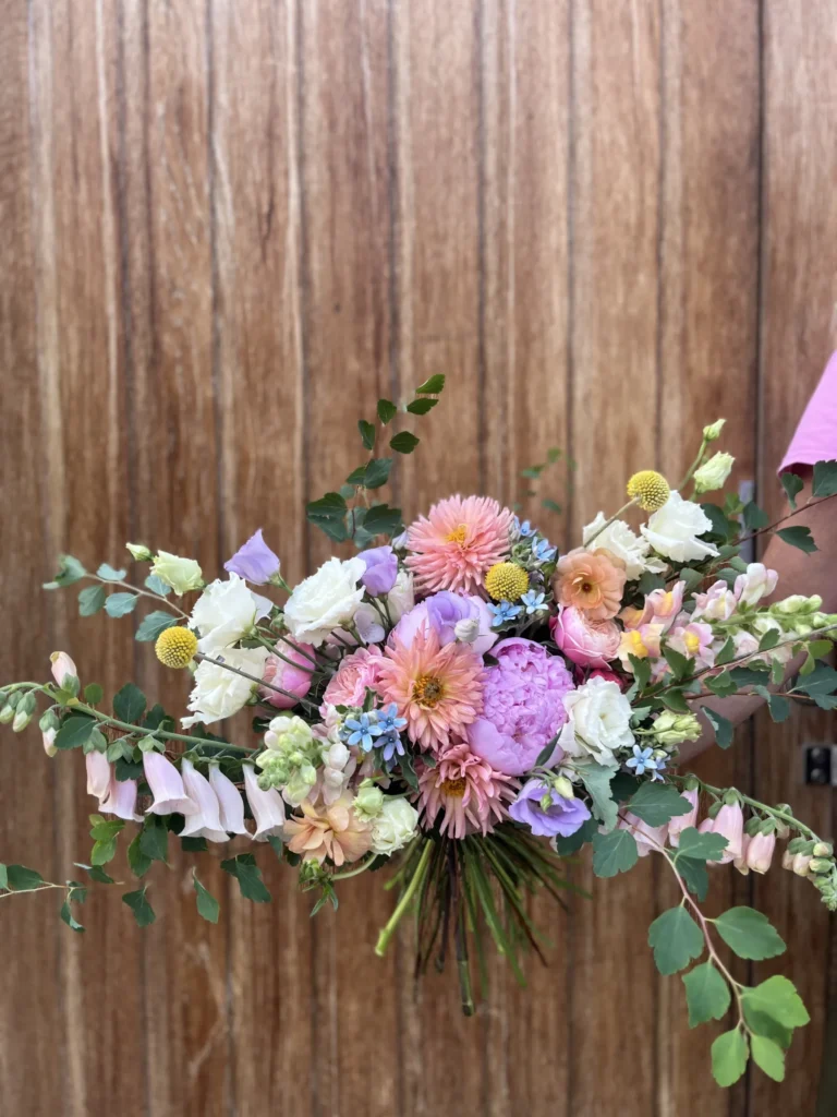 En hand håller en bukett med olika blommor, inklusive rosa dahlior, vita rosor, gul craspedia och grönska, framför en träbakgrund.