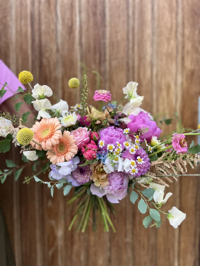 En färgglad bukett med blandade blommor, inklusive rosa, persika, lila och gula blommor med gröna blad, hålls framför en träbakgrund.