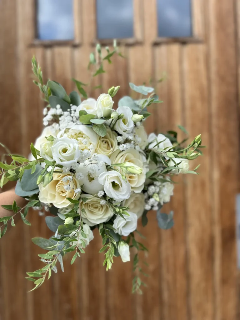 En hand håller en bukett med vita och gräddfärgade rosor, babys breath och grönska framför en trädörr.
