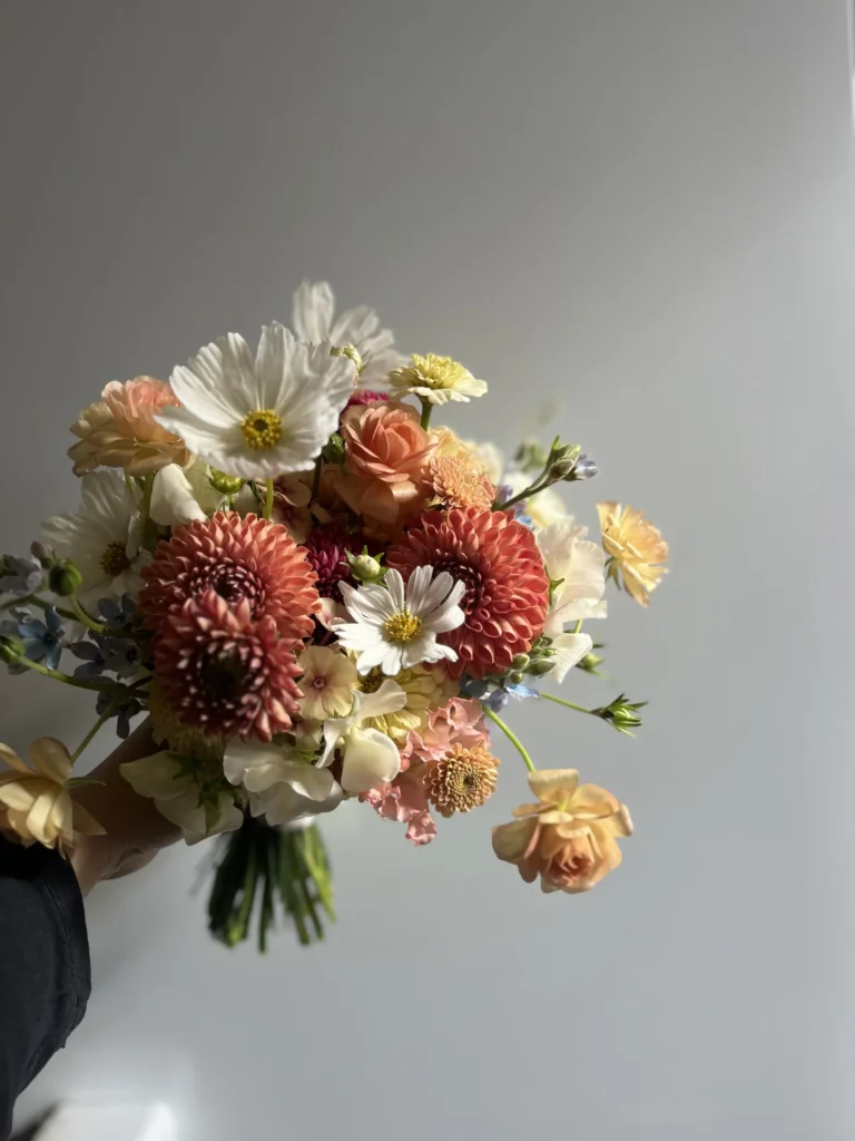 En hand håller i en bukett med olika blommor, bland annat dahlior, prästkragar och rosor, mot en grå bakgrund.