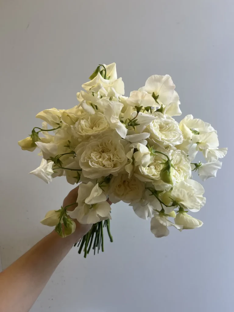 En hand håller i en bukett gräddfärgade blommor med gröna stjälkar mot en ljusgrå bakgrund.