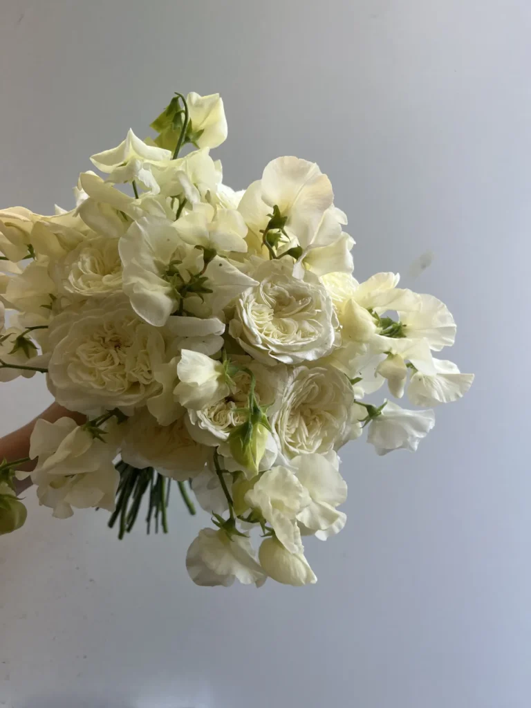 En hand håller i en bukett med gräddfärgade rosor och luktärtsblommor mot en ljusgrå bakgrund.