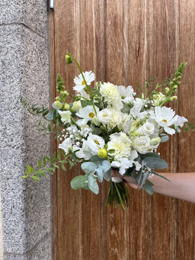 En hand håller i en bukett med vita blommor och grönska framför en trädörr och en stenmur.