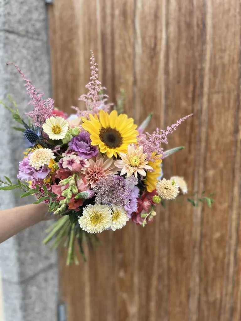 En hand håller en färgglad bukett med blandade blommor, inklusive solrosor, krysantemum och asters, framför en trädörr.
