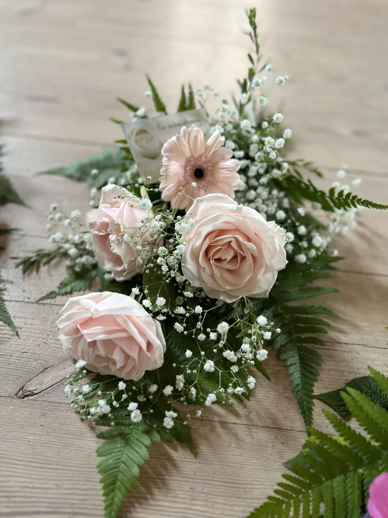 Ett blomsterarrangemang med ljusrosa rosor, babys breath, en enda rosa gerbera och gröna ormbunkar på en träyta.