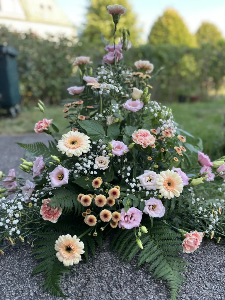 Ett blomsterarrangemang med rosa, persikofärgade och gräddfärgade blommor, ormbunkar och grönska utomhus på en asfalterad yta med en trädgårdsbakgrund.