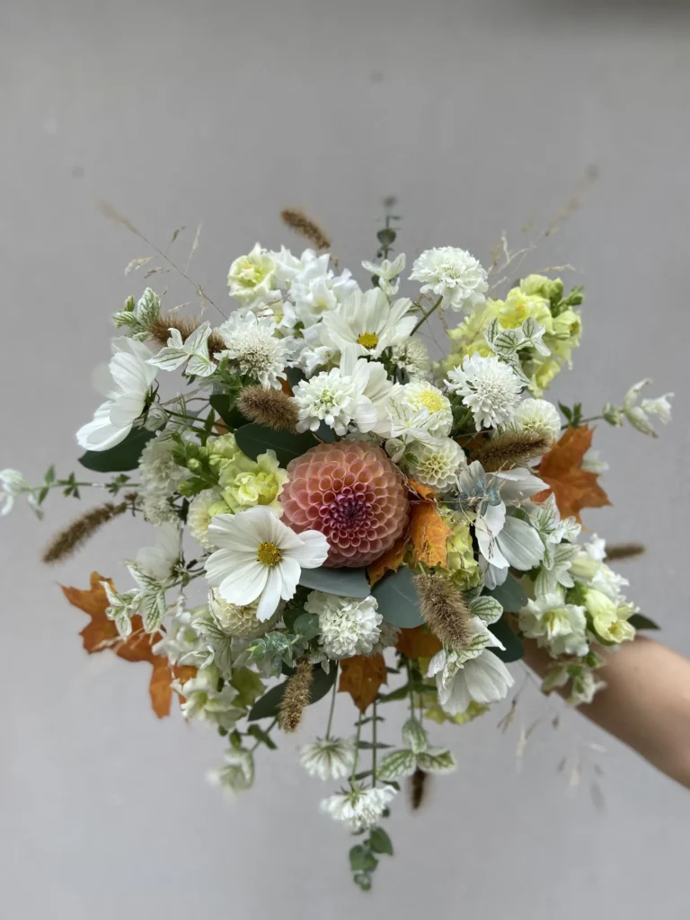 En hand håller i en bukett med vita och gula blommor, en rosa dahlia i mitten, gröna blad, brunt gräs och orange höstlöv mot en neutral bakgrund.