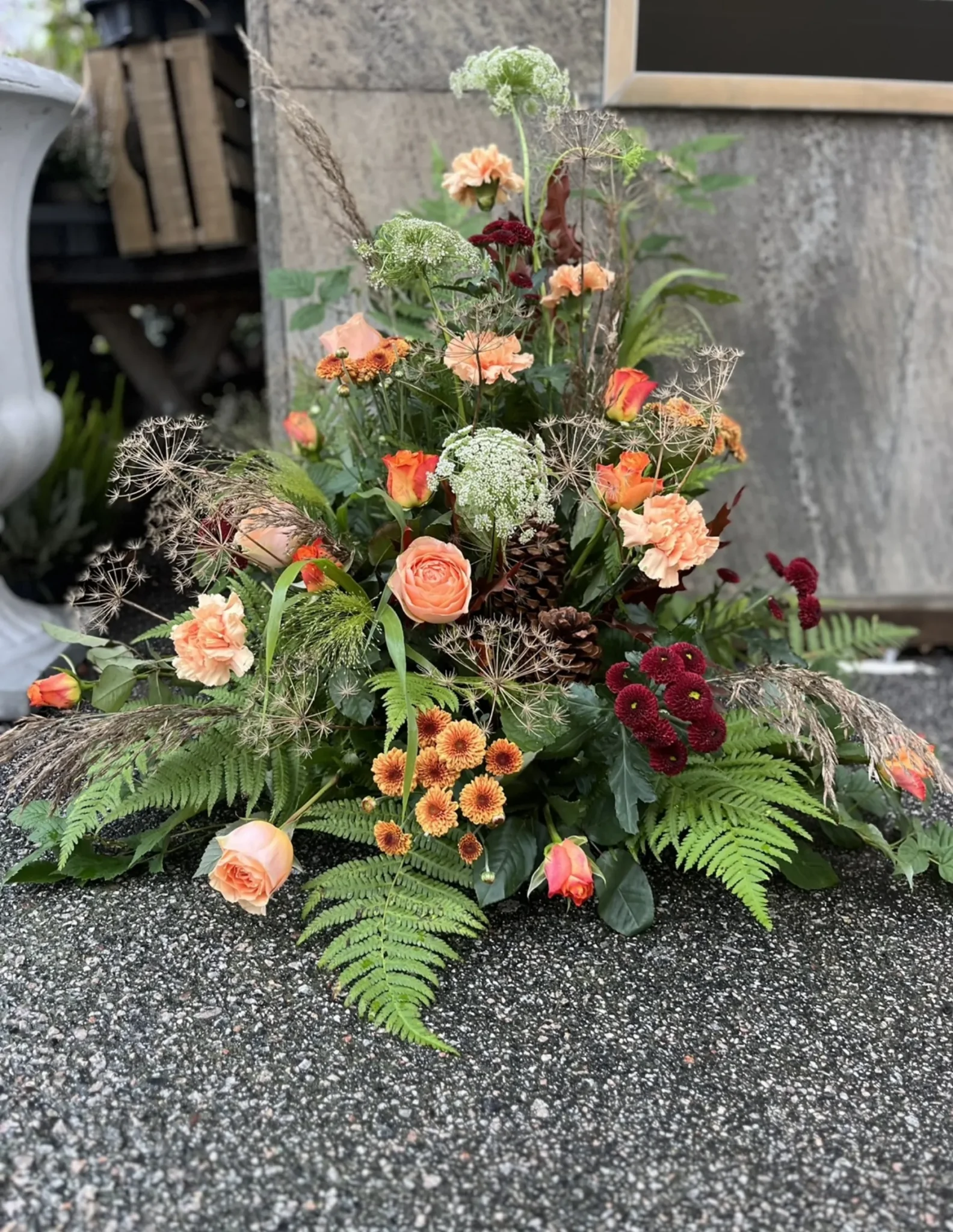 Ett blomsterarrangemang med orangefärgade rosor, mörkröda blommor, ormbunkar och grönska, placerat utomhus på en grusyta.