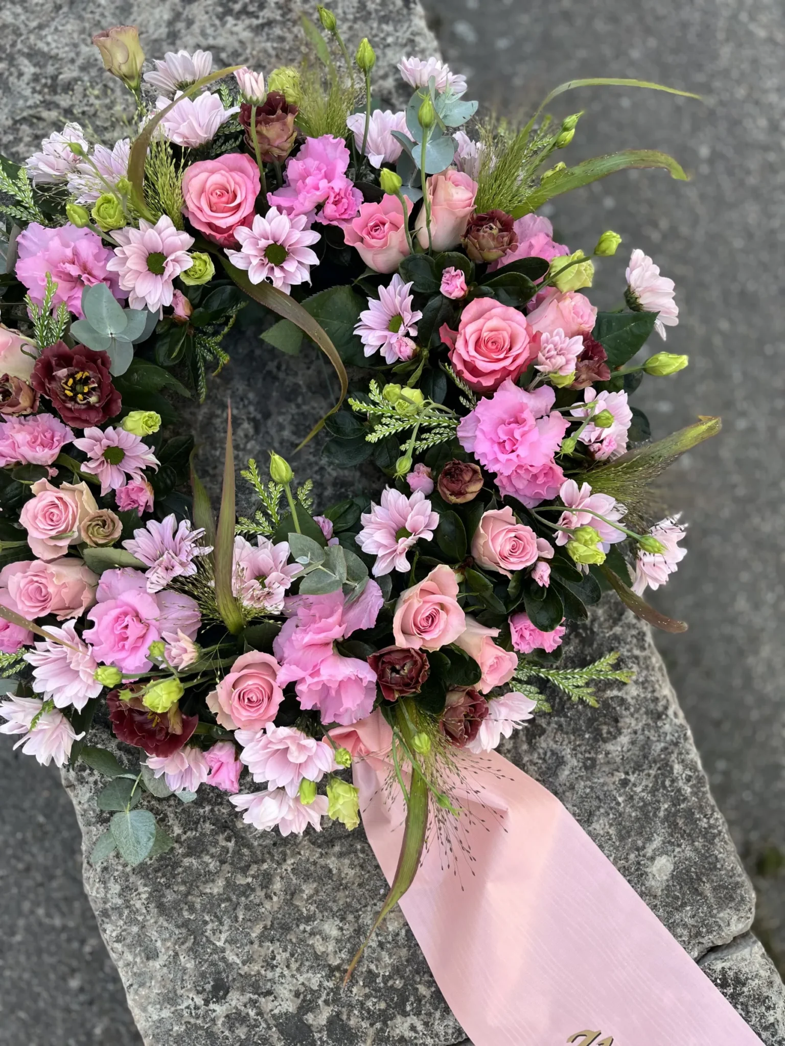 En blomsterkrans med rosa och vinröda blommor, grönska och ett rosa band är placerad på en grå stenyta.