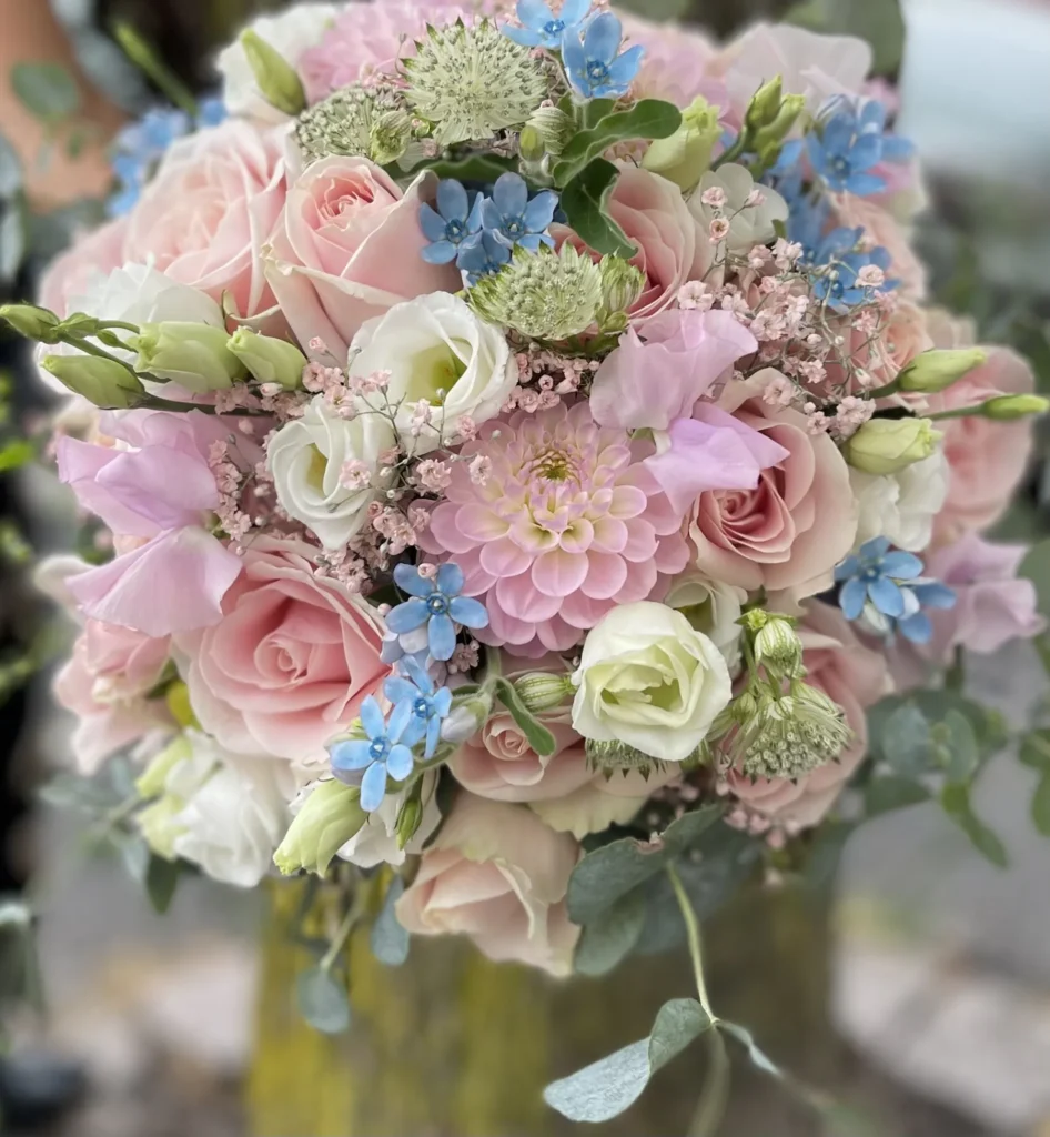 En bukett med rosa rosor, vita blommor, blå blommor och grönska arrangerad i en rund, kompakt stil.