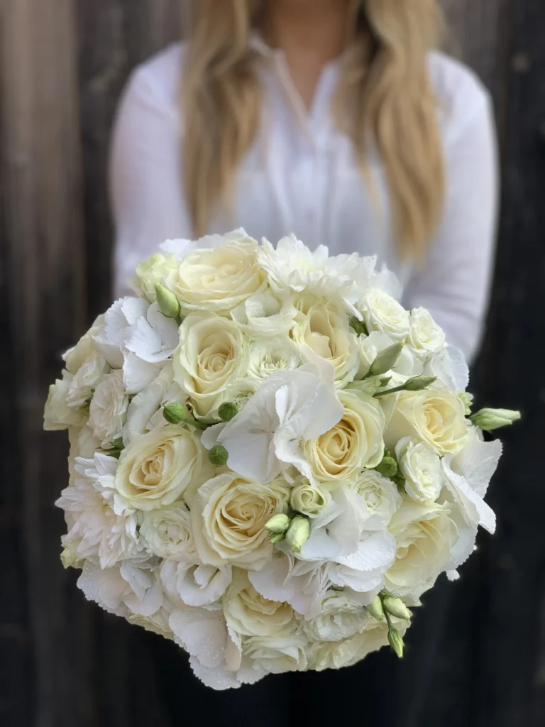 En person i vit skjorta håller en bukett med vita och gräddfärgade rosor, lisianthus och hortensior mot en mörk träbakgrund.