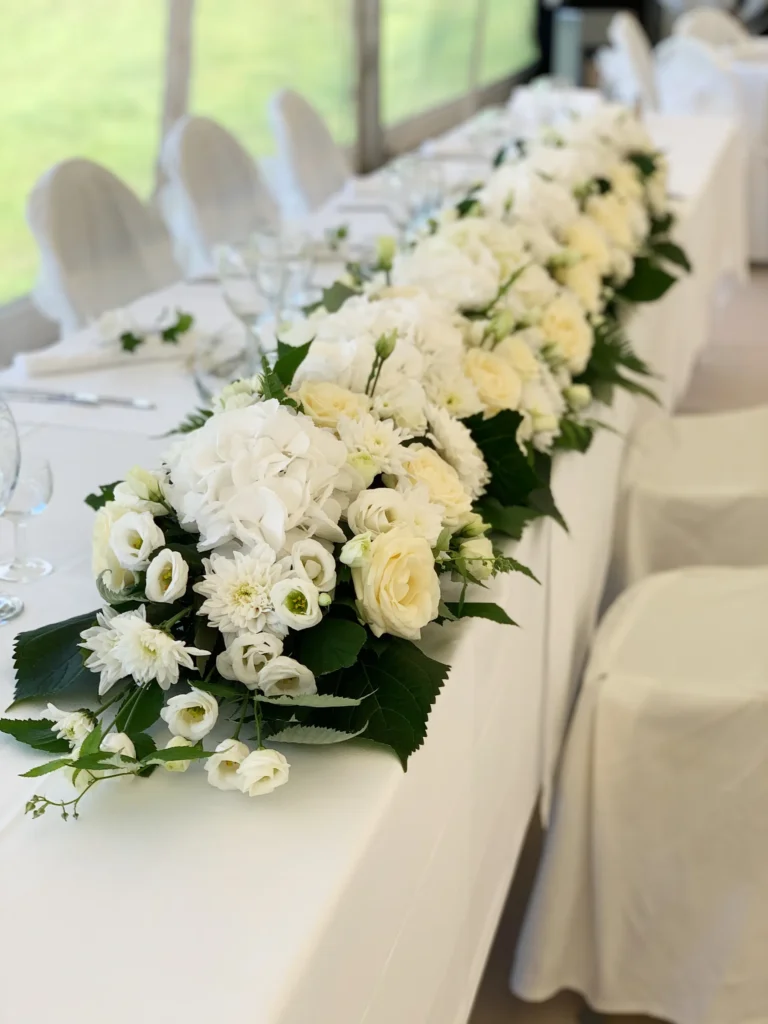 Ett långbord med vit duk och stolar, dekorerat med en mittpunkt av vita och gräddfärgade blommor och gröna blad, dukat för en formell tillställning.