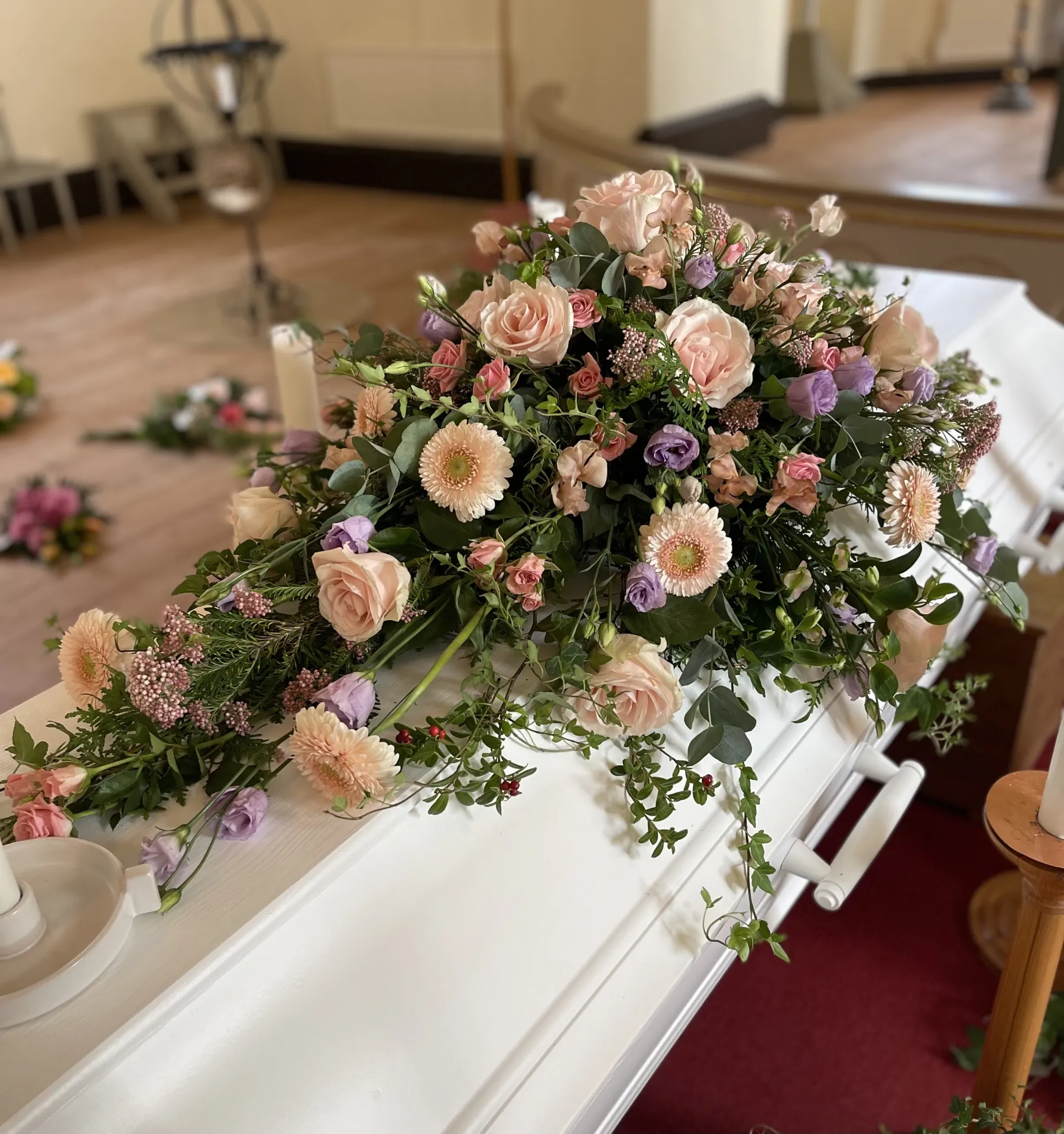 En vit kista med ett stort arrangemang av rosa och lila blommor ovanpå, placerad i ett rum med trägolv och utspridda blomsterhyllningar.