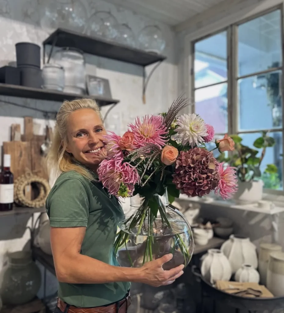 En kvinna står inomhus och håller i en stor glasvas fylld med färgglada blommor och ler mot kameran. Hyllor med köksutrustning och ett fönster syns i bakgrunden.