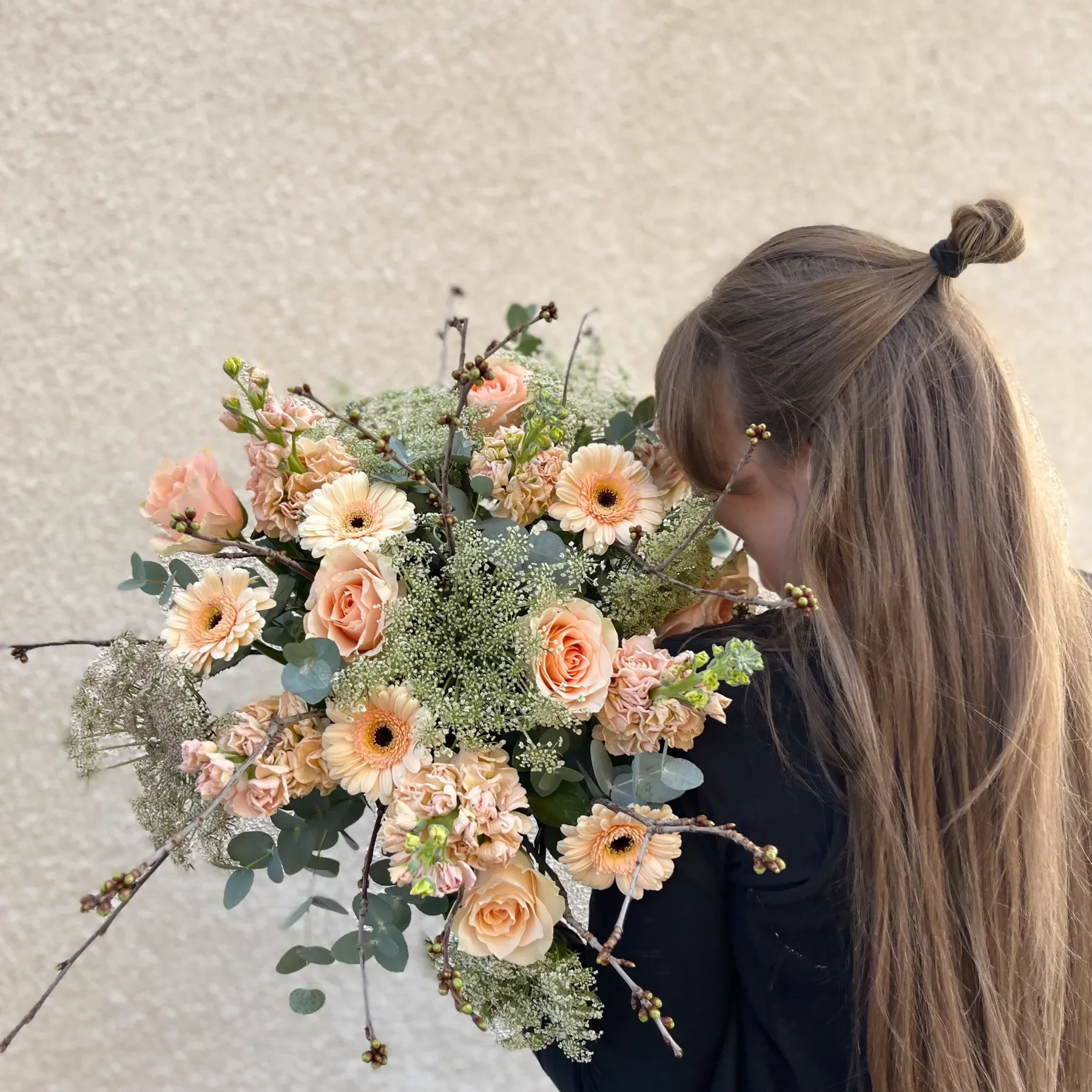 Brudbukett En person med långt brunt hår i en halvuppsatt knut håller en stor bukett med persikoblommor och grönska mot en beige texturerad vägg.