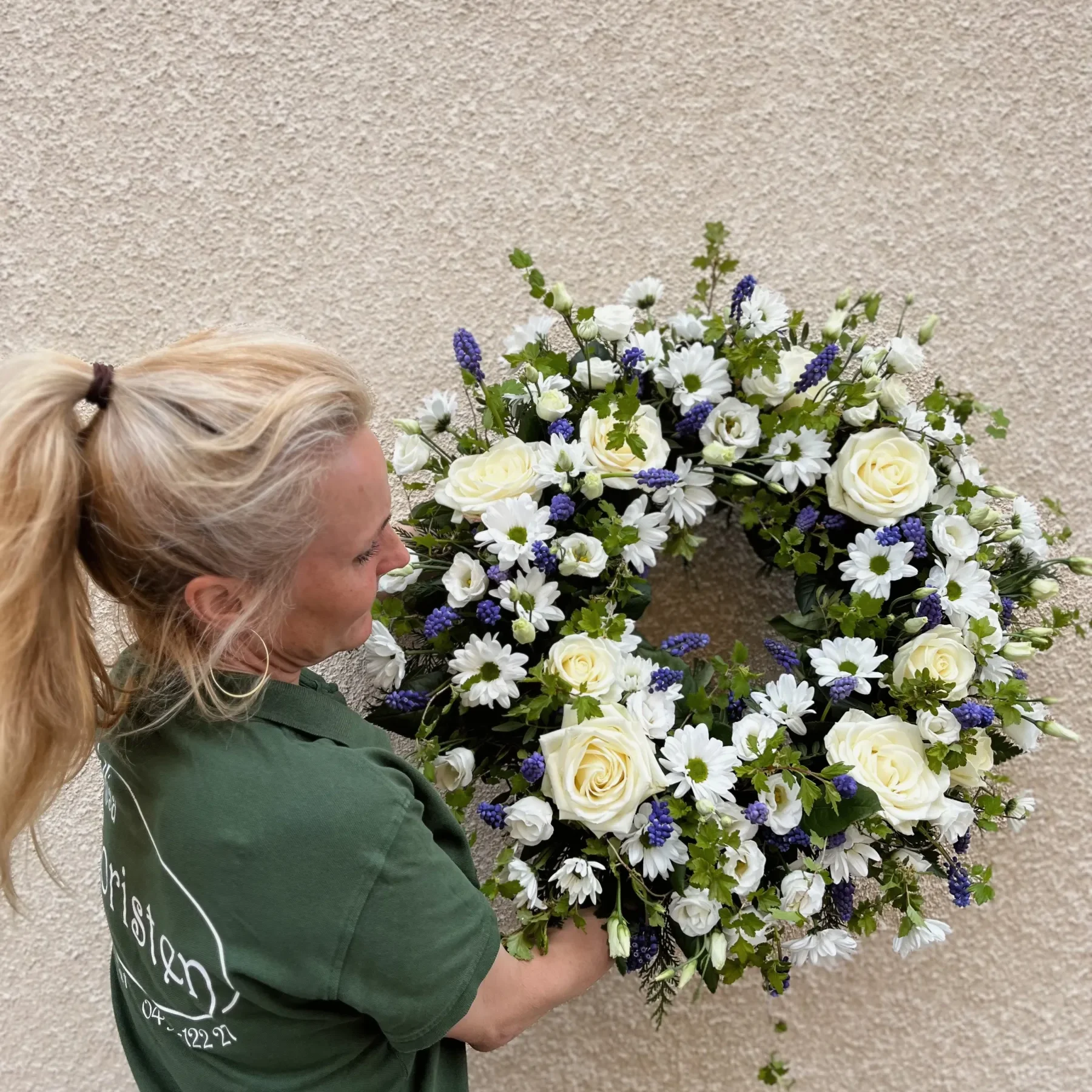 Begravning kransar16 En person i grön skjorta håller en stor rund blomsterkrans med vita och lila blommor mot en ljus strukturerad vägg.