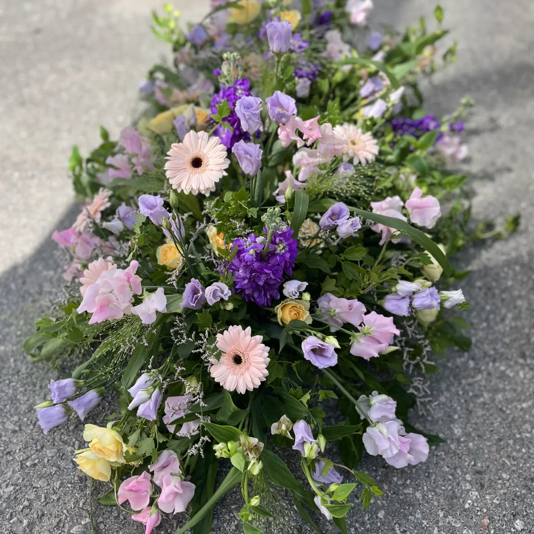 Ett blomsterarrangemang med rosa, lila och gula blommor, inklusive gerberor och lisianthus, placerat på en grå utomhusyta.