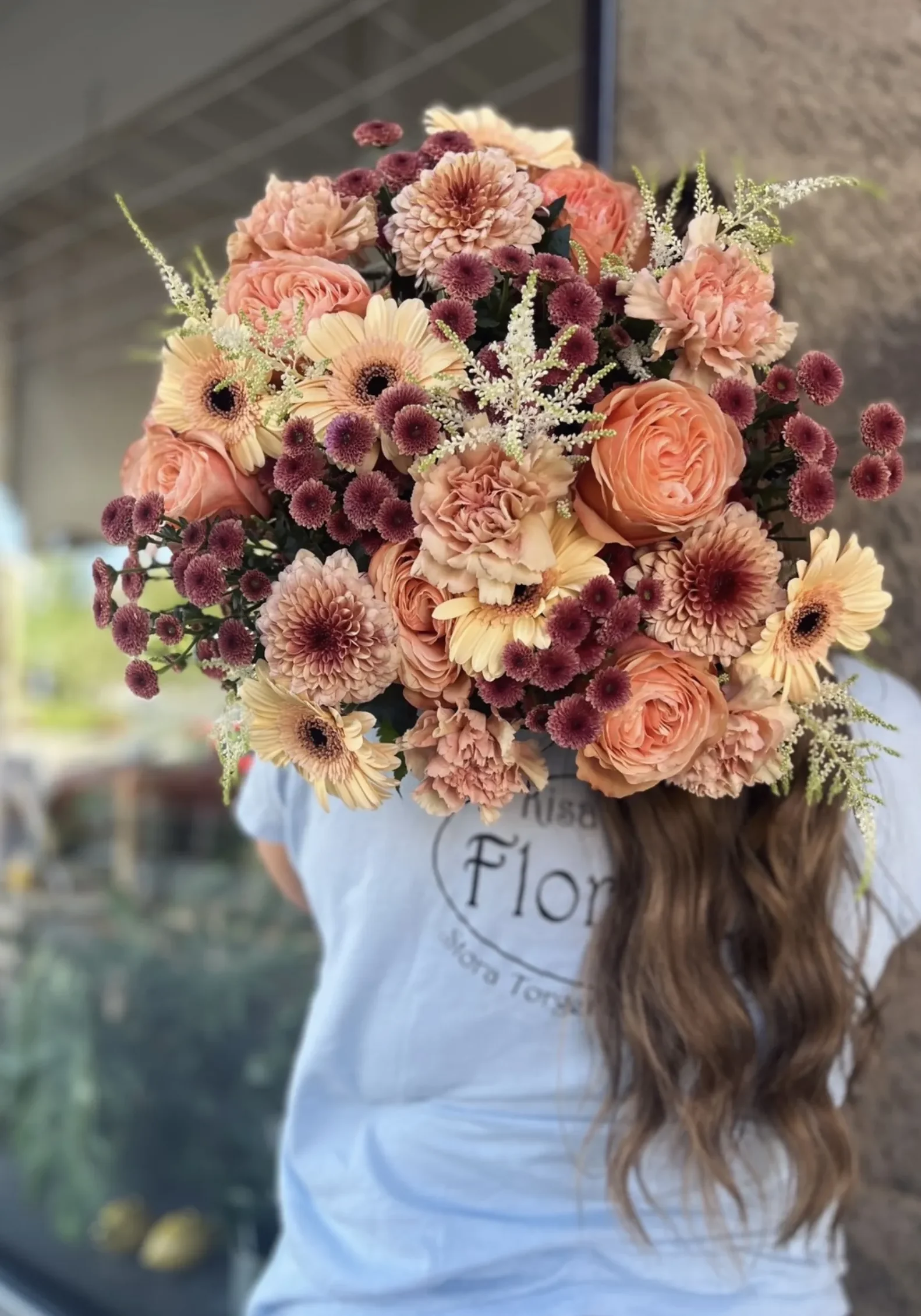 Vardagsbukett En person med långt brunt hår står utanför en byggnad och håller i en stor bukett med persikofärgade och vinröda blommor som döljer ansiktet.
