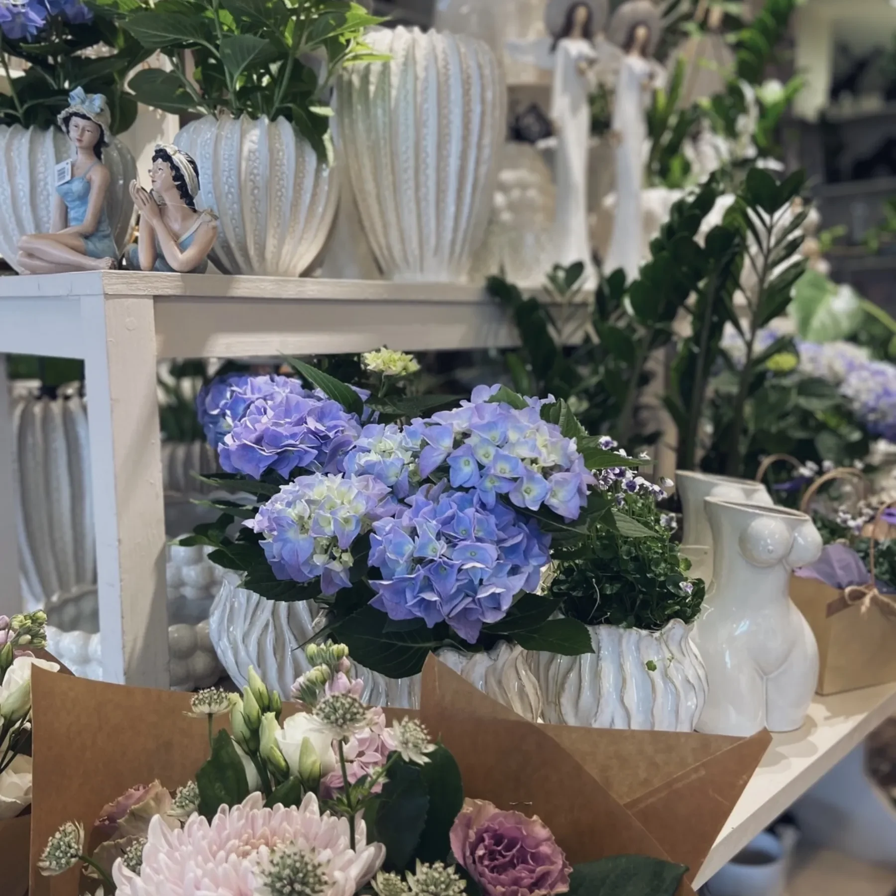 Butiken Blå hortensior och diverse blommor i vita keramikvaser, omgivna av dekorativa figurer och växter på vita hyllor.