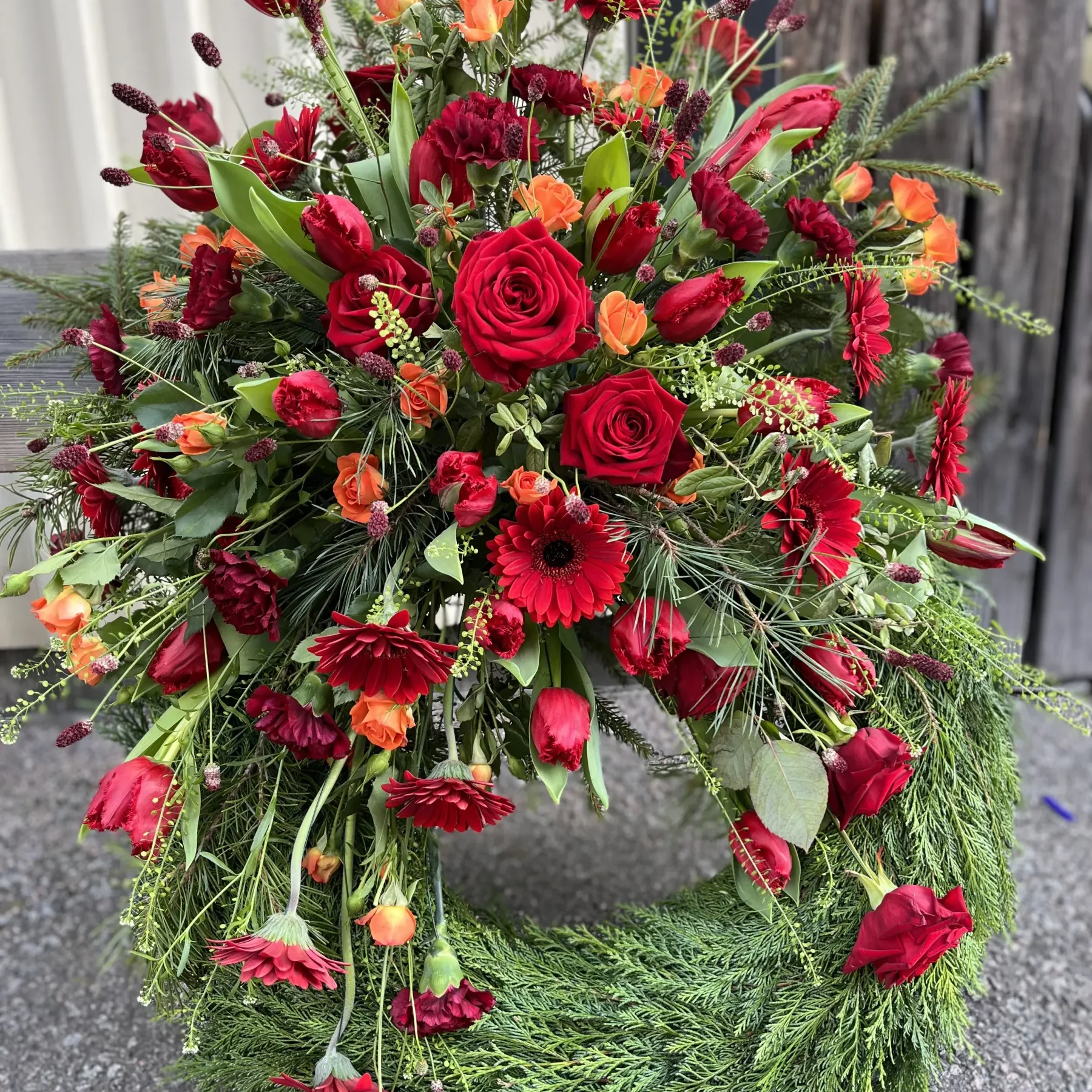En krans dekorerad med röda rosor, gerbera daisy, tulpaner, orange rosor och grönska, placerad på en betongyta mot en bakgrund av korrugerad plåt och trä.