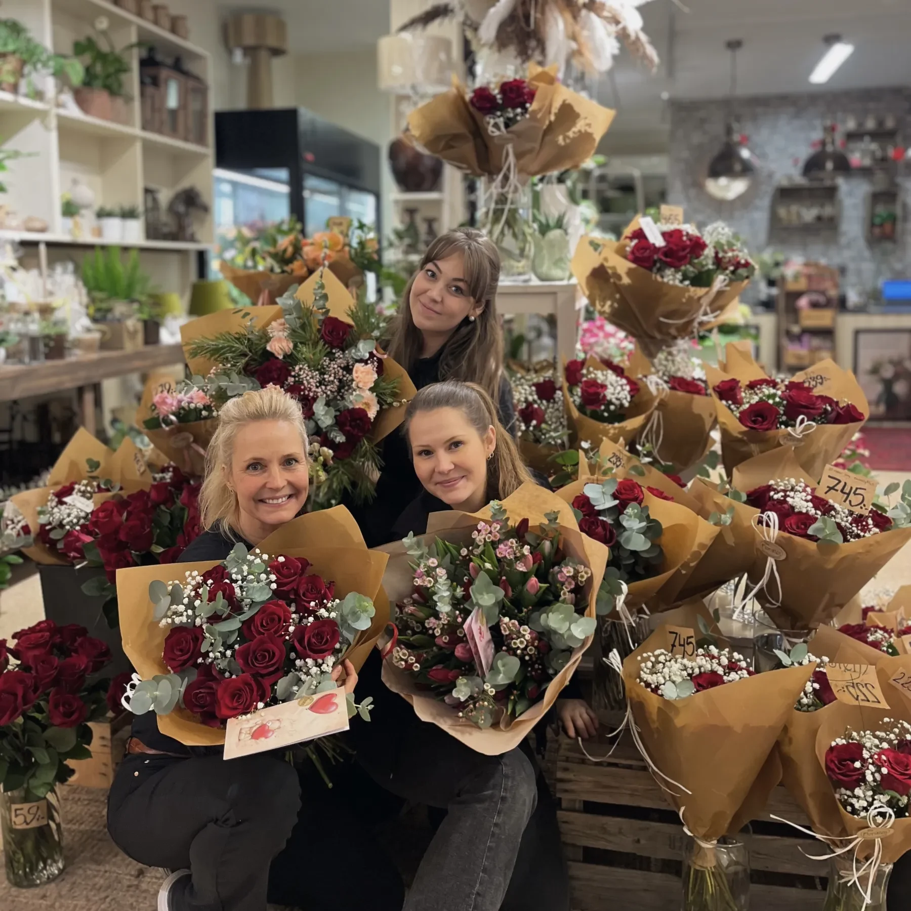 Kisafloristen Tre kvinnor poserar i en blomsteraffär, omgivna av buketter med röda och vita blommor inslagna i brunt papper. I bakgrunden syns olika växter och blomsterarrangemang.