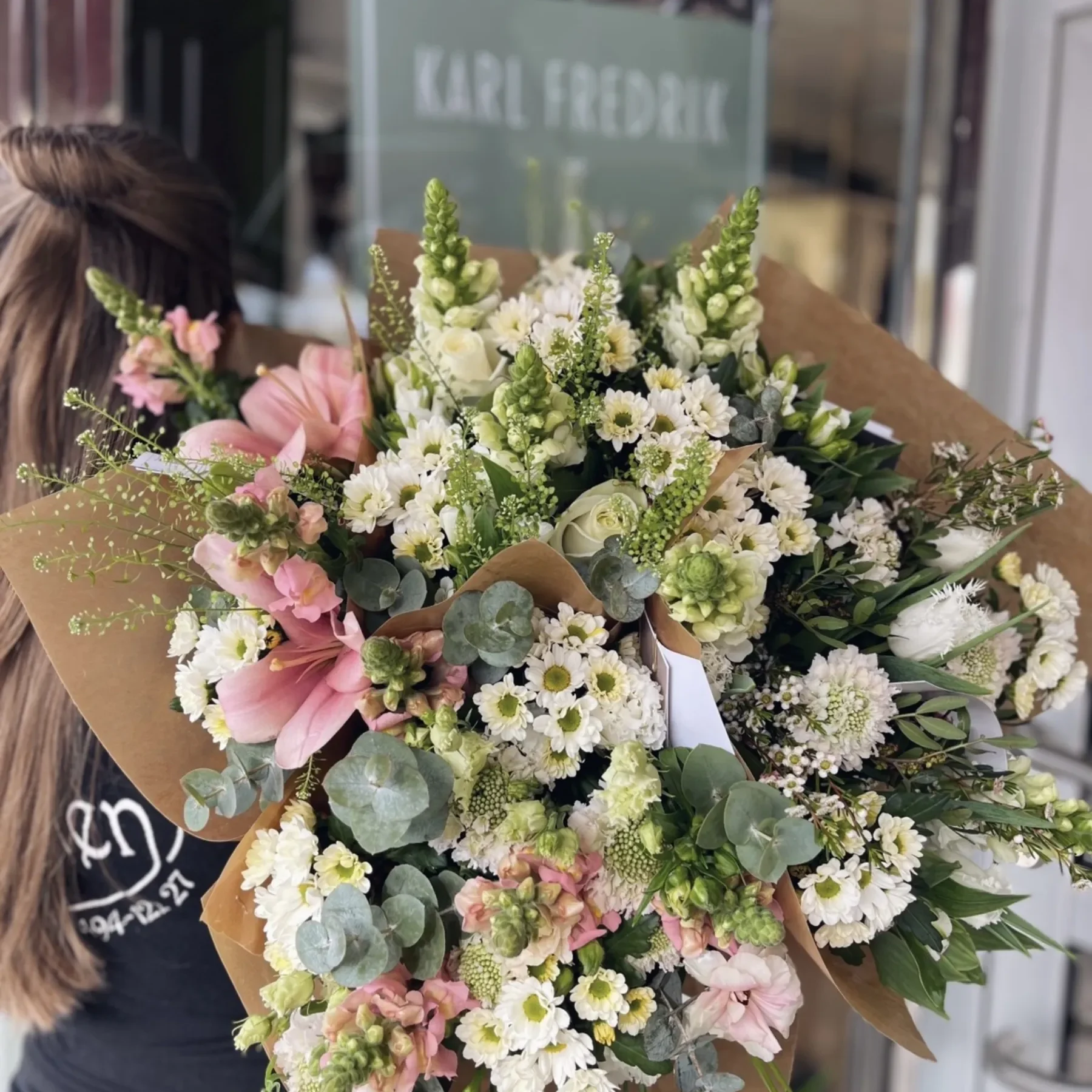 En person håller i en stor bukett med blandade blommor, bland annat vita, rosa och gröna, framför en butiksentré med texten KARL FREDRIK på glaset.